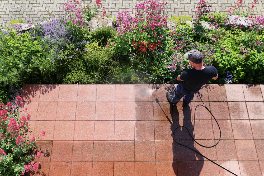 person pressure washing a red tile patio near a garden full of flowers